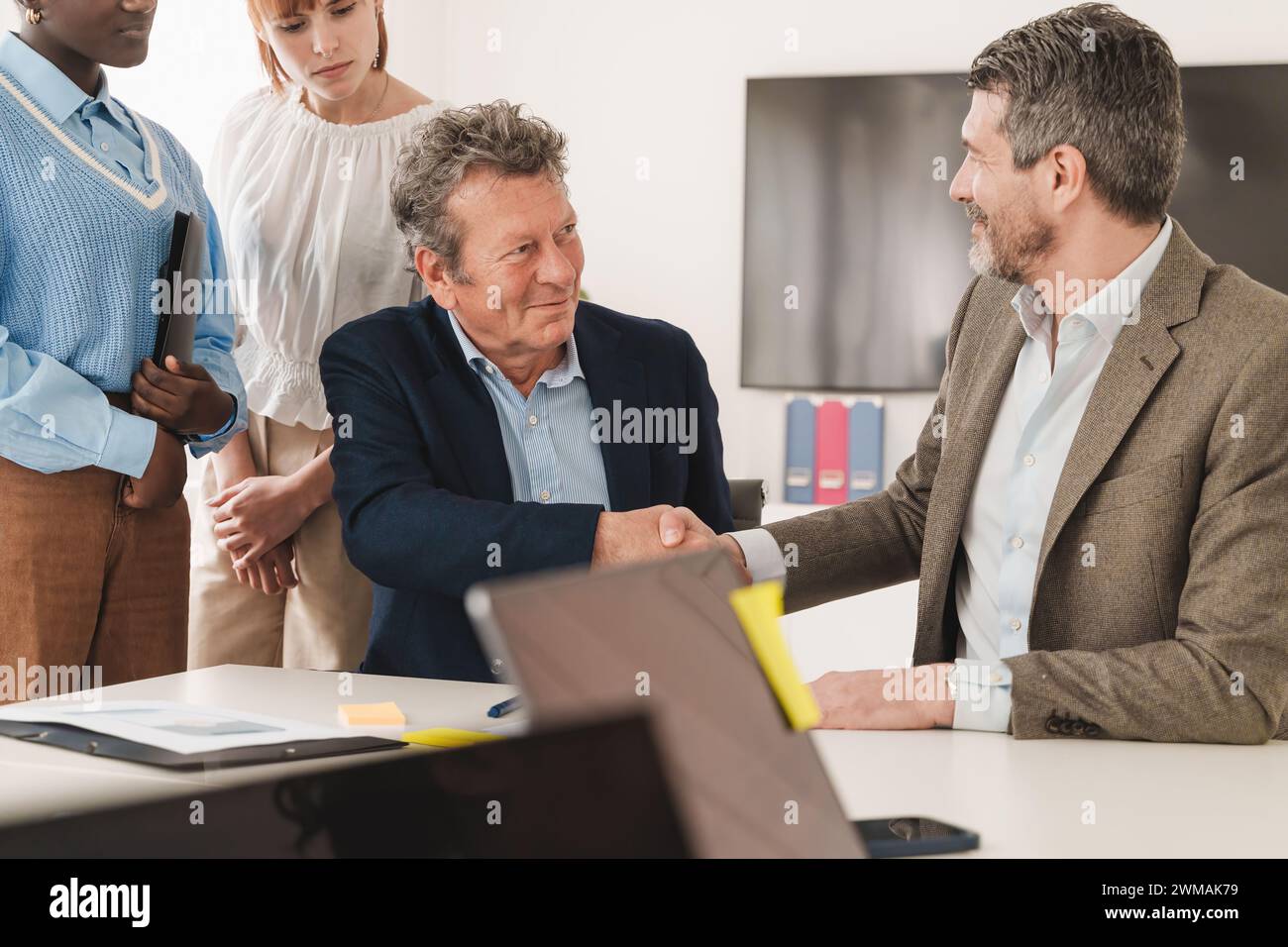 Due uomini d'affari che stringono la mano in ufficio con il team watching - accordo e concetto di partnership con colleghi multietnici Foto Stock