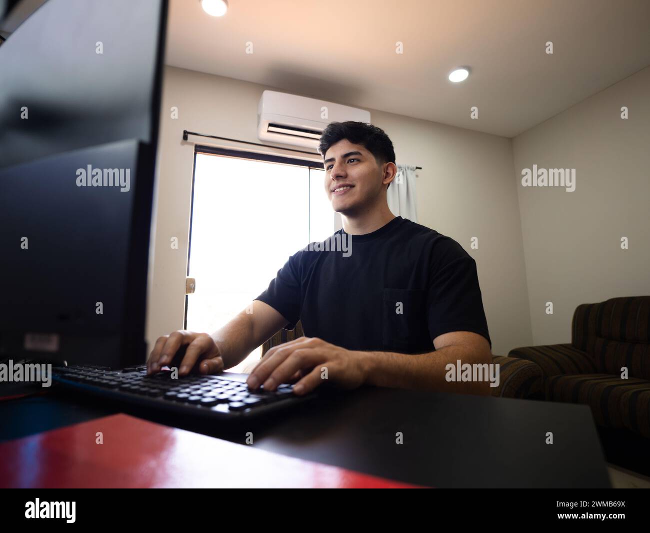 Un giovane sorridente e dedicato davanti al computer, che scrive incarichi, progetta progetti e si immerge nel mondo del lavoro a distanza Foto Stock