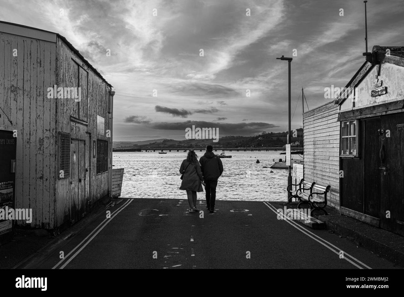 Giovane coppia che cammina verso il fiume Teign al tramonto. Cielo spettacolare con il fiume Teign e il ponte Shaldon in lontananza. Foto in bianco e nero. Foto Stock