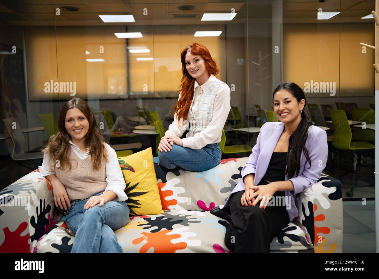 Tre donne professionali sedute casualmente su un vivace divano a motivi geometrici in un ufficio creativo. Foto Stock