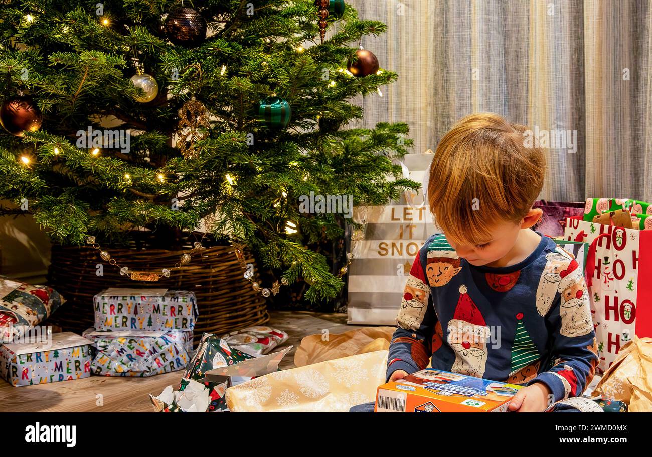 Bambino di 5 anni che ispeziona un Natale appena aperto presente davanti all'albero con pj con Babbo Natale ed Elfo su regali non aperti intorno a lui Foto Stock