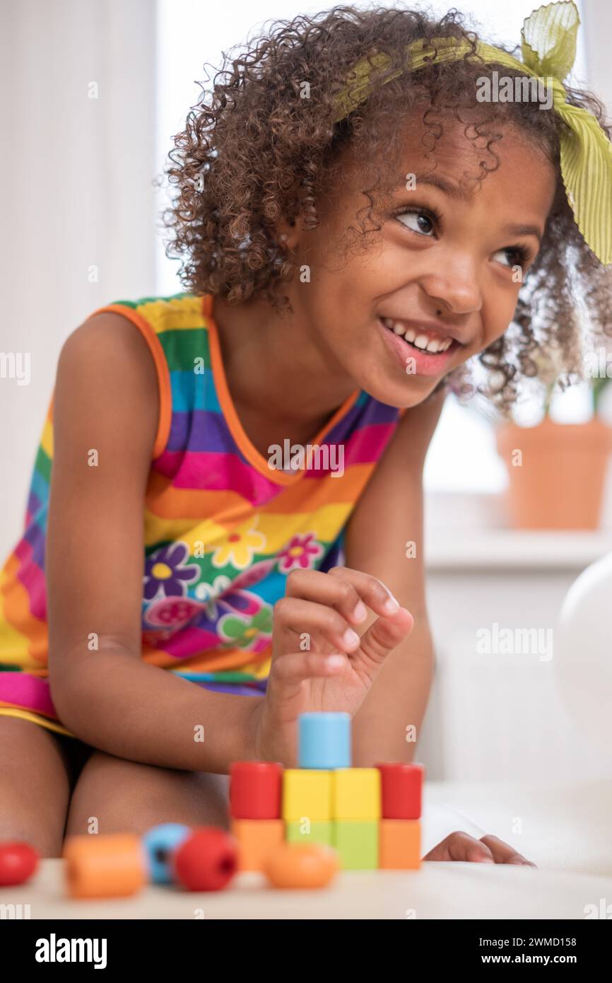 Ragazza afroamericana sorridente dai capelli ricci con fiocco giallo sorride mentre si impilano blocchi, vivace abito a righe arcobaleno. per contenuti sulla gioia di apprendere Foto Stock