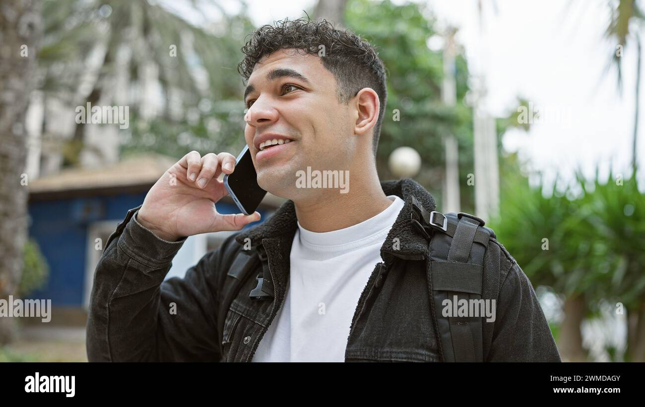 Un bell'uomo con uno zaino che sorride mentre parla al telefono cellulare in una strada cittadina. Foto Stock