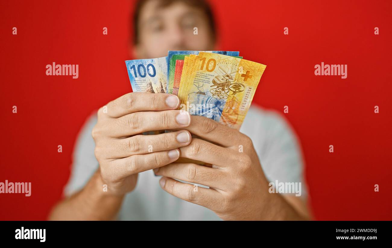 Giovane, biondo, barbuto, bell'uomo che banca grandi e alte puntate che contano banconote in franchi svizzeri! isolato su un impressionante sfondo rosso Foto Stock