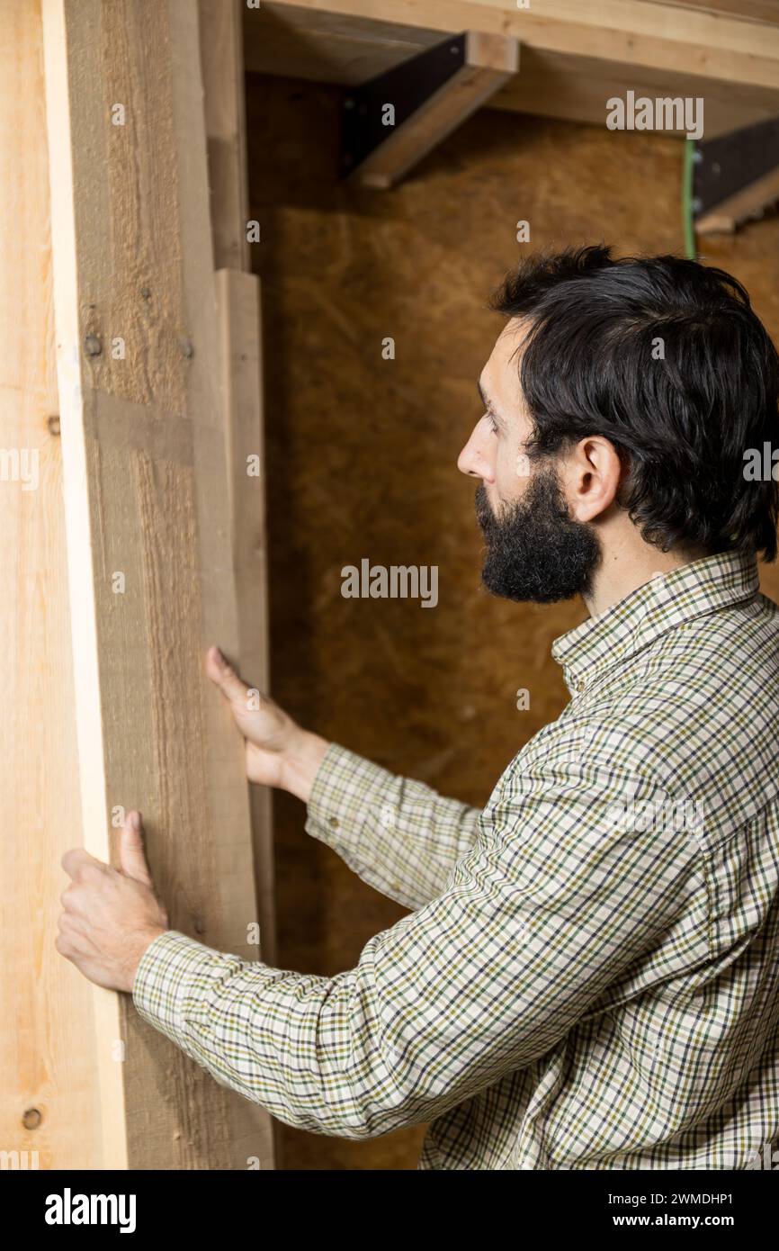 Il falegname concentrato con foto verticale smista le tavole di legno, selezionando il pezzo perfetto per il suo prossimo progetto in un deposito ben organizzato Foto Stock