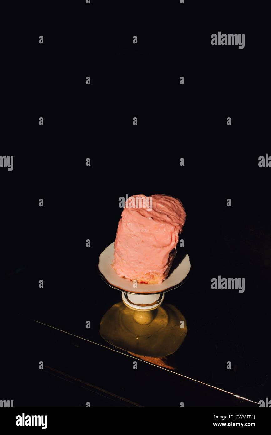 piccola torta a forma di cuore alta e pendente con glassa rosa pallido su piedistallo in ceramica bianca su un tavolo dorato con sfondo nero Foto Stock