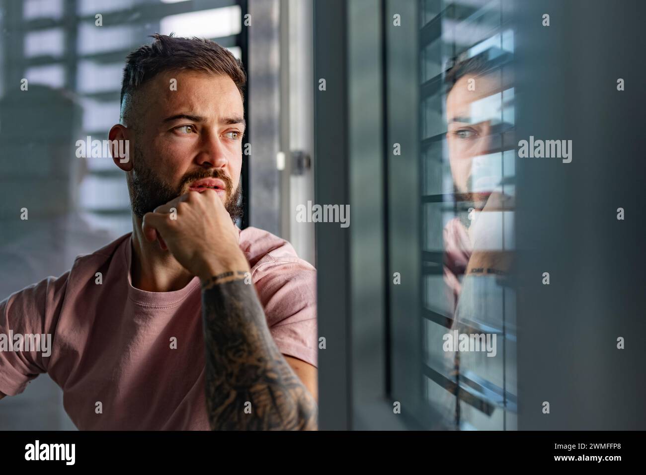 Bell'uomo calmo in maglietta rosa con tatuaggio sulle braccia che guarda dalla finestra. Foto Stock
