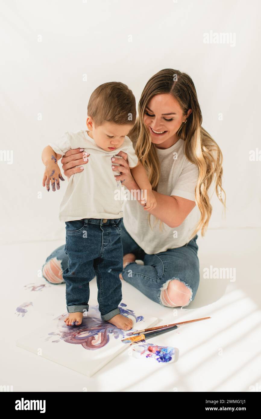 La madre sorridente aiuta il figlio a piedi nudi a fare impronte con la vernice su tela Foto Stock