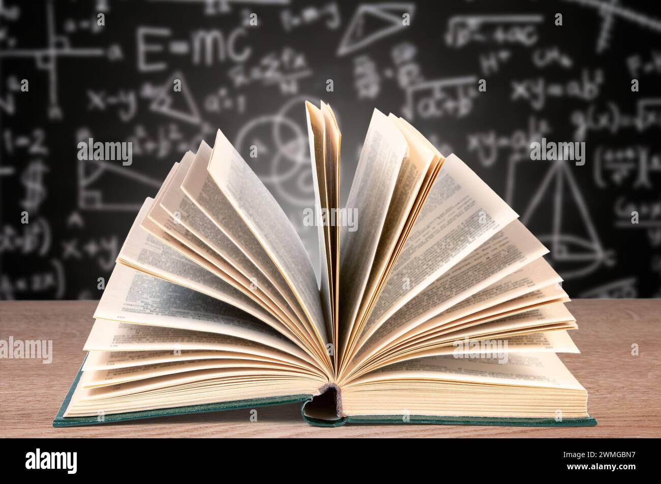 Un libro di testo aperto giace su una scrivania di legno in primo piano con uno sfondo sfocato caratterizzato da una lavagna coperta da varie equazioni matematiche Foto Stock