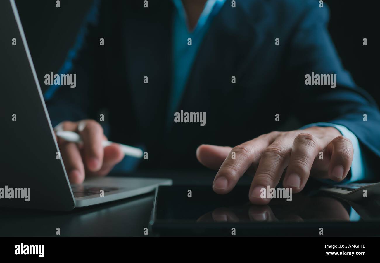 Uomo d'affari in tuta nera che lavora su computer portatile e tablet, mani a sfioramento su tablet in ufficio con sfondo scuro, lavoro online, primo piano, Foto Stock