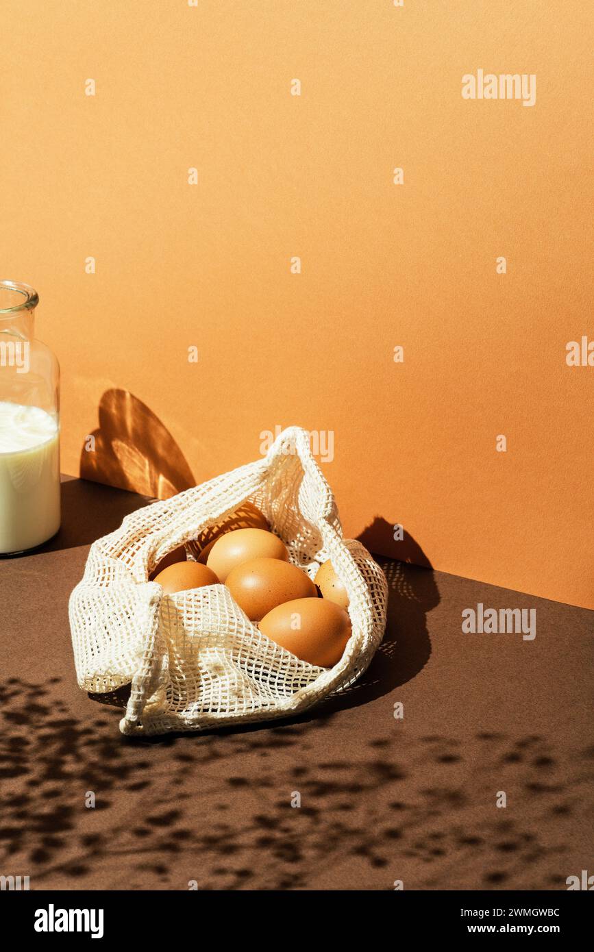 Uova in un sacchetto in rete con latte nel biberon di vetro su sfondo marrone. Effetto luce solare con ombre floreali e scatti ad angolo alto. Copia sfondo spazio. Foto Stock