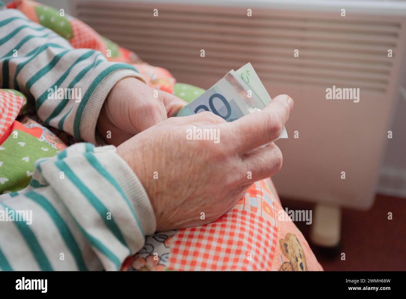 Primo piano delle mani. Un'anziana donna caucasica siede su una sedia sotto una coperta con un riscaldatore vicino ai piedi e tiene i soldi nelle sue mani. Prezzi in aumento Foto Stock