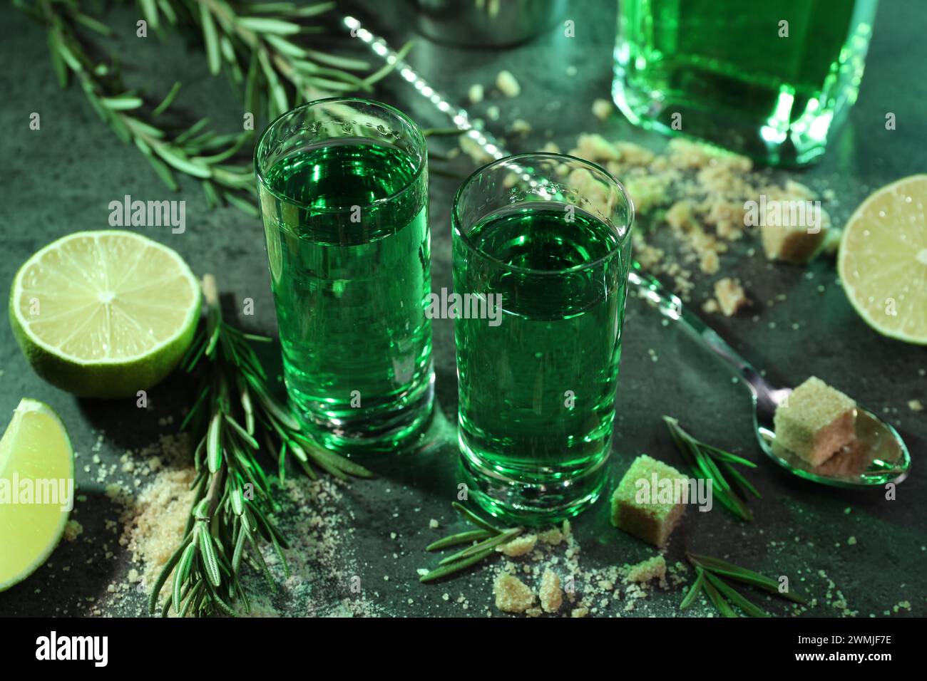 Absinthe in bicchieri da shot, cucchiaio, zucchero di canna, lime e rosmarino su un tavolo grigio, primo piano. Bevanda alcolica Foto Stock