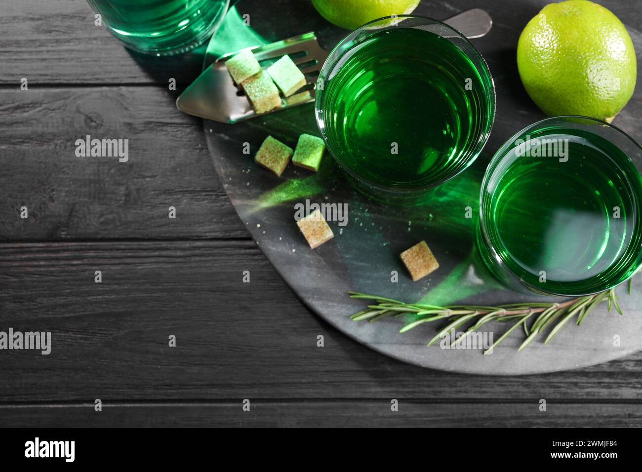 Absinthe in bicchieri, rosmarino, zucchero di canna e lime su un tavolo in legno nero, vista dall'alto con spazio per il testo. Bevanda alcolica Foto Stock