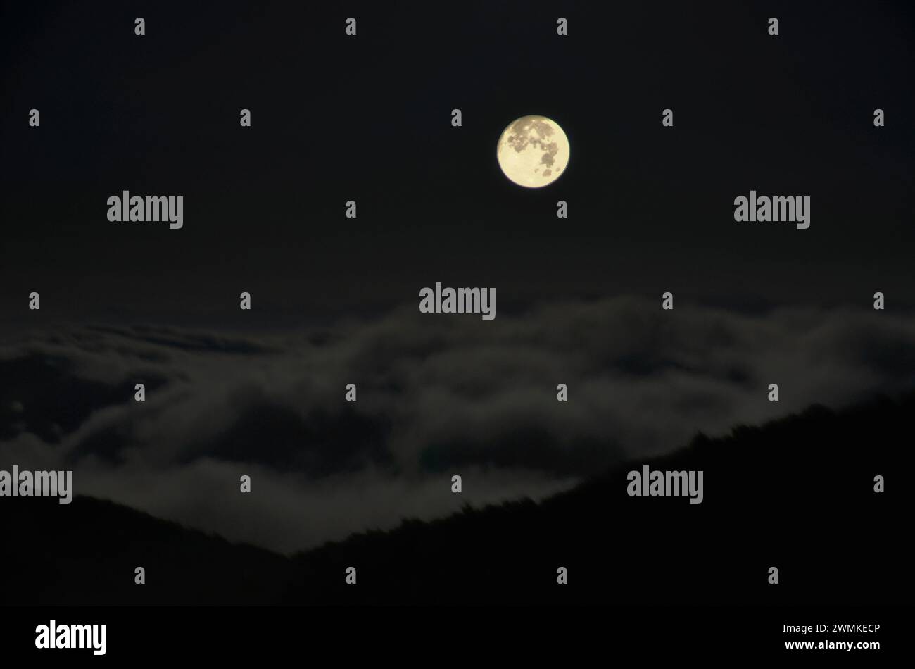 La luna piena si illumina nel cielo buio di notte con nuvole sulle montagne sagomate Foto Stock