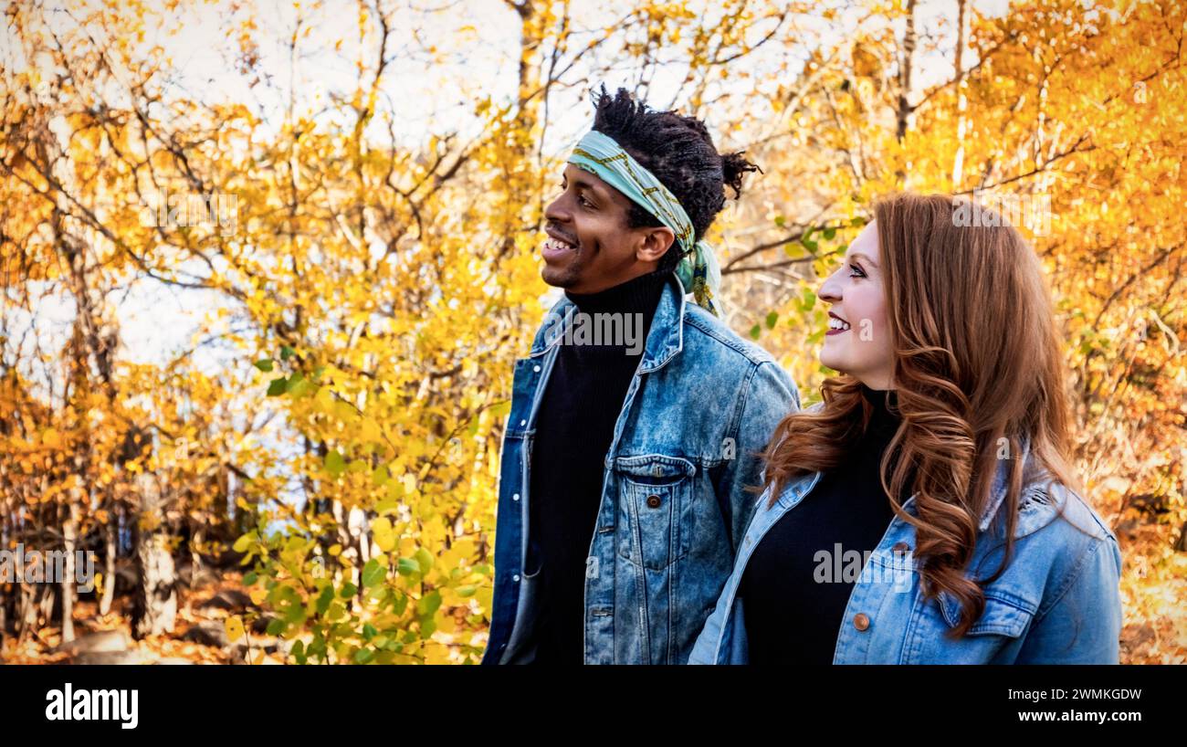 Coppia di sposi con corse miste, sorridenti mentre camminano attraverso un parco cittadino durante un'uscita di famiglia autunnale, trascorrendo del tempo insieme Foto Stock