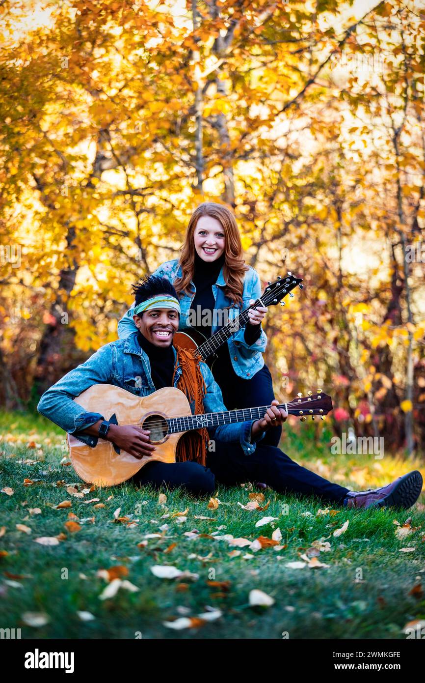 Ritratto di una coppia sposata di razza mista, sorridente e in posa per la telecamera che tiene le chitarre mentre si siede sull'erba durante un'uscita di famiglia autunnale io... Foto Stock