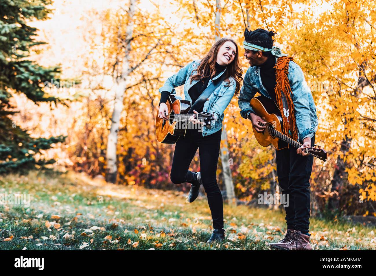 Coppia di sposi in gara mista che trascorrono del tempo insieme, guardandosi sorridendo e suonando le chitarre durante una gita in famiglia autunnale in un parco cittadino Foto Stock