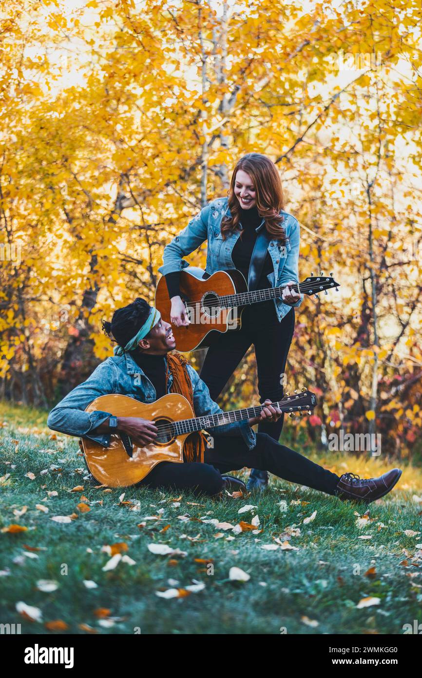 Coppia di sposi in gara mista che trascorrono del tempo insieme, sorridendo e suonando le chitarre durante una gita in famiglia autunnale in un parco cittadino Foto Stock