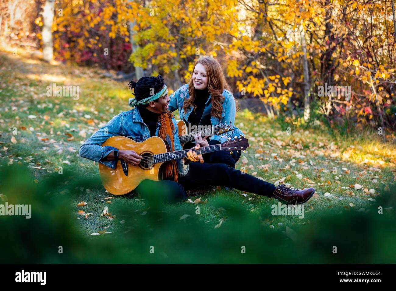 Guarda attraverso le foglie di una coppia sposata di razza mista seduta sull'erba, trascorrendo del tempo insieme, sorridendo e suonando le chitarre durante un fami autunnale... Foto Stock