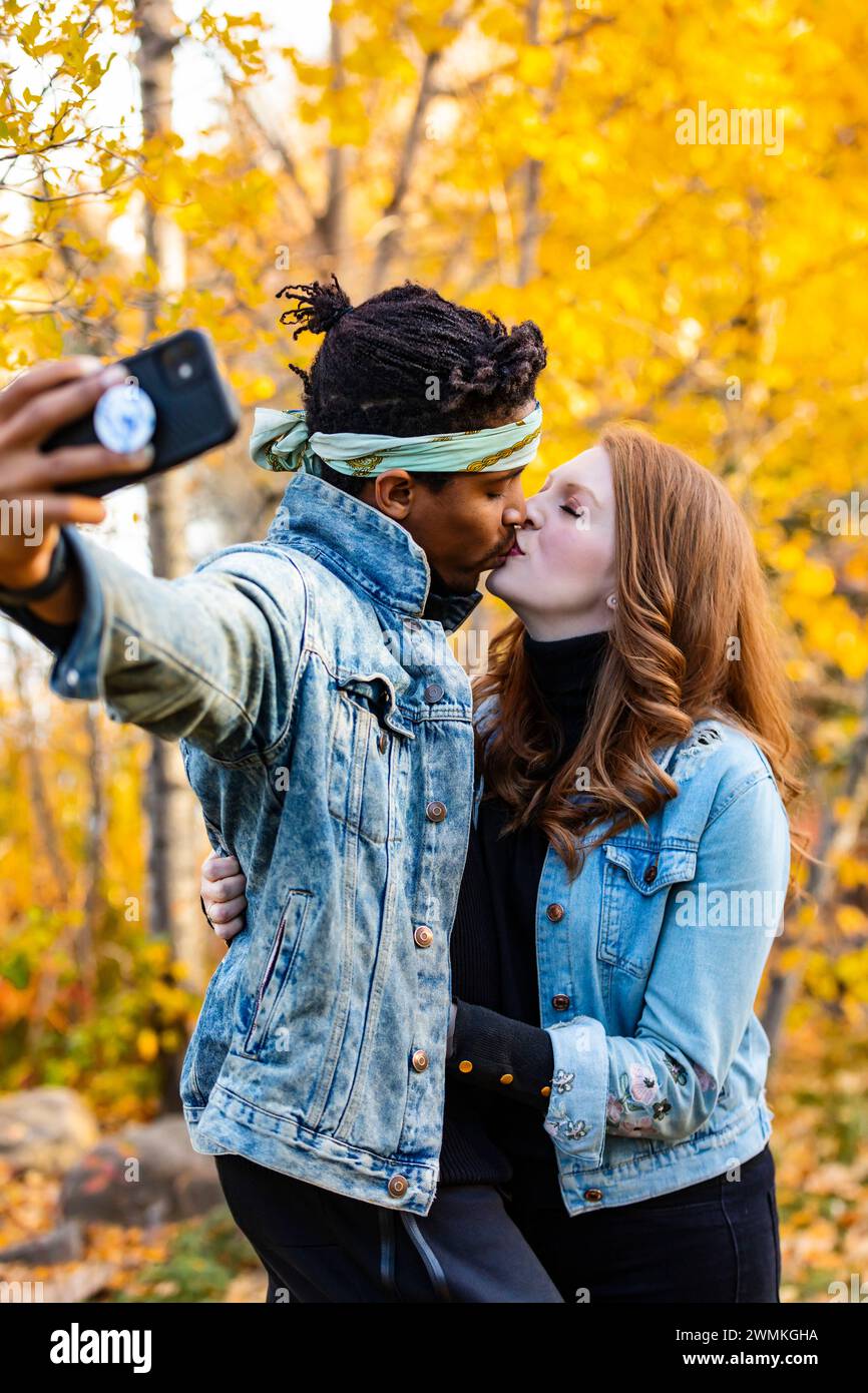 Primo piano di una coppia di corse miste che si baciano con un selfie, trascorrendo del tempo insieme durante una gita in famiglia autunnale in un parco cittadino Foto Stock