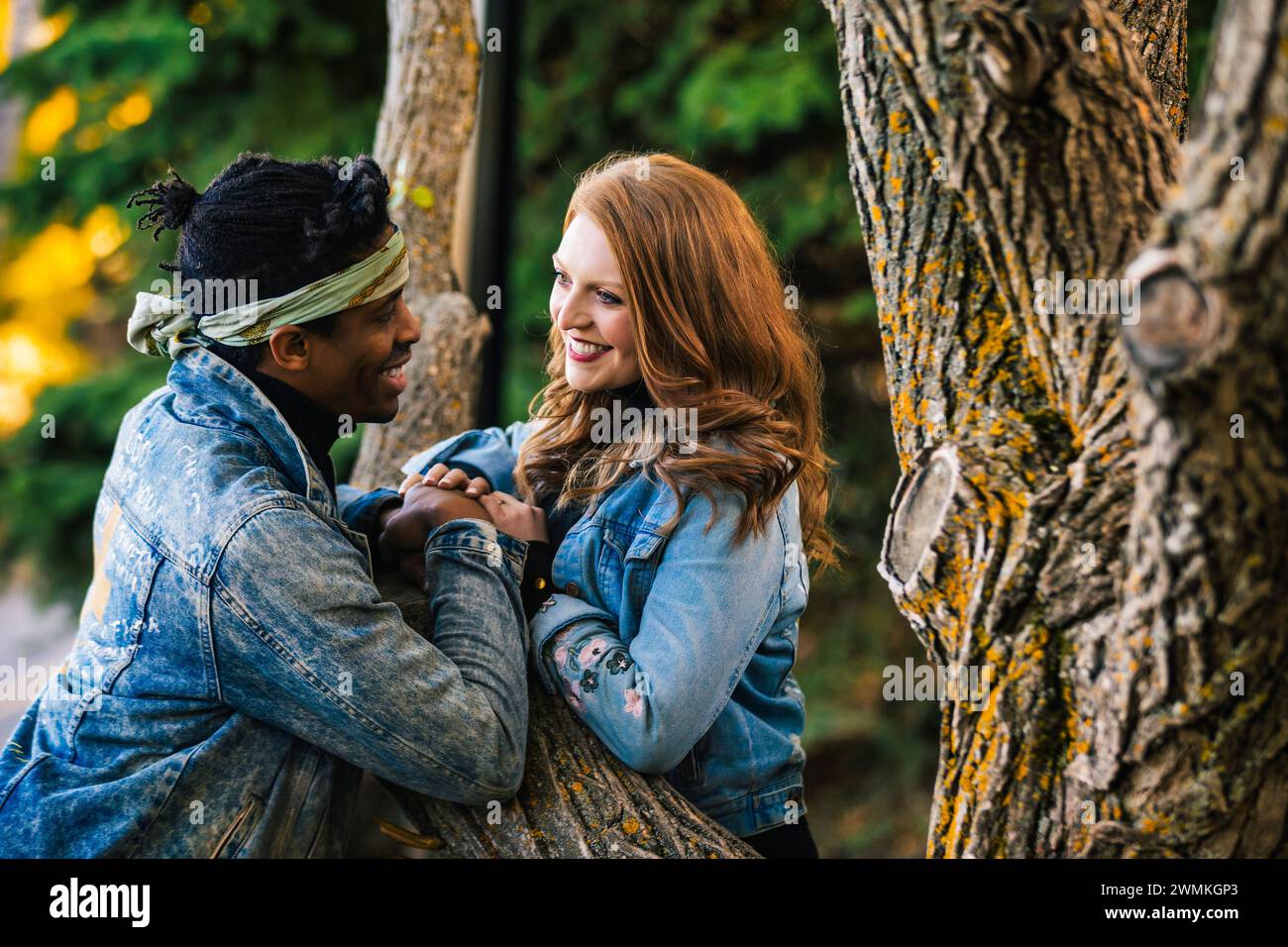 Primo piano di una coppia mista che sorride e si tiene per mano mentre si riposa su un ramo d'albero, trascorrendo del tempo insieme durante un fal... Foto Stock
