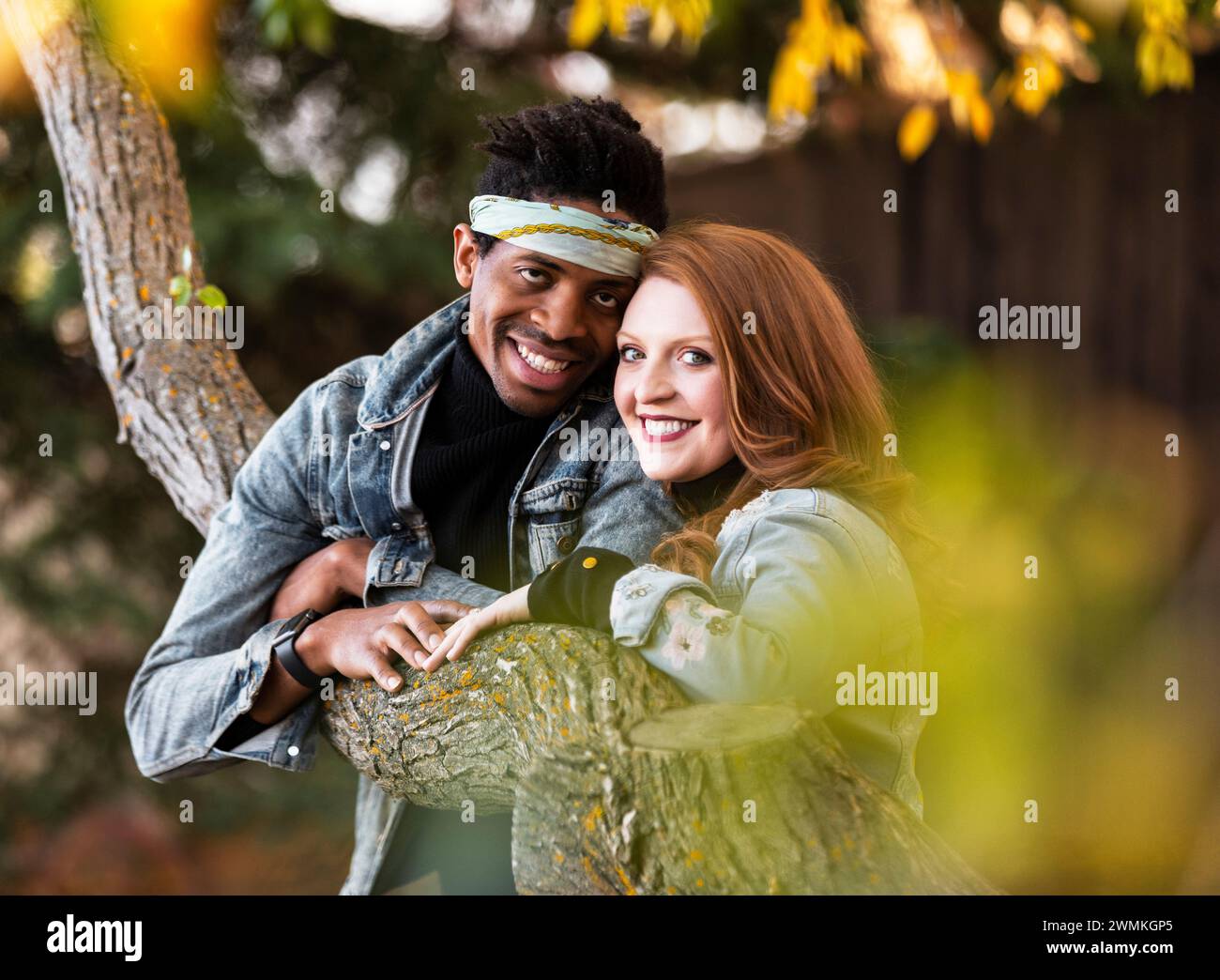 Ritratto ravvicinato di una coppia di razze miste sorridente e tenuta per mano mentre si riposa su un ramo d'albero, trascorrendo del tempo insieme durante una fam autunnale... Foto Stock