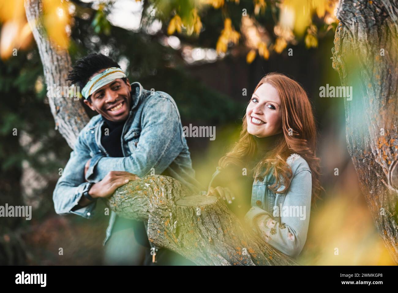 Primo piano di una coppia di corse miste sorridente e godendosi la vita all'aria aperta mentre si riposa su un ramo d'albero, trascorrendo del tempo insieme durante un fami autunnale... Foto Stock