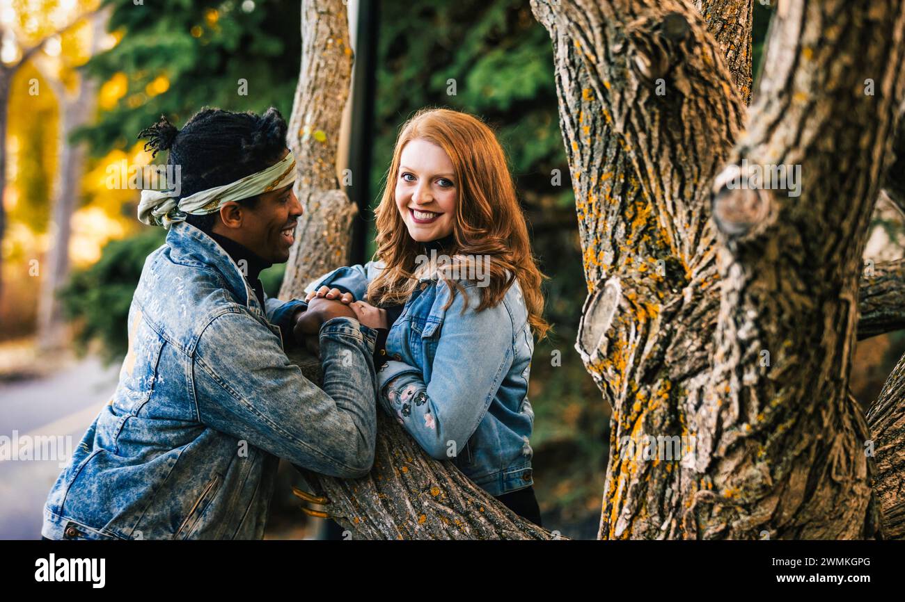 Primo piano di una coppia mista sorridente e tenuta per mano mentre riposa su un ramo d'albero, trascorrendo del tempo insieme durante un'uscita di famiglia autunnale... Foto Stock