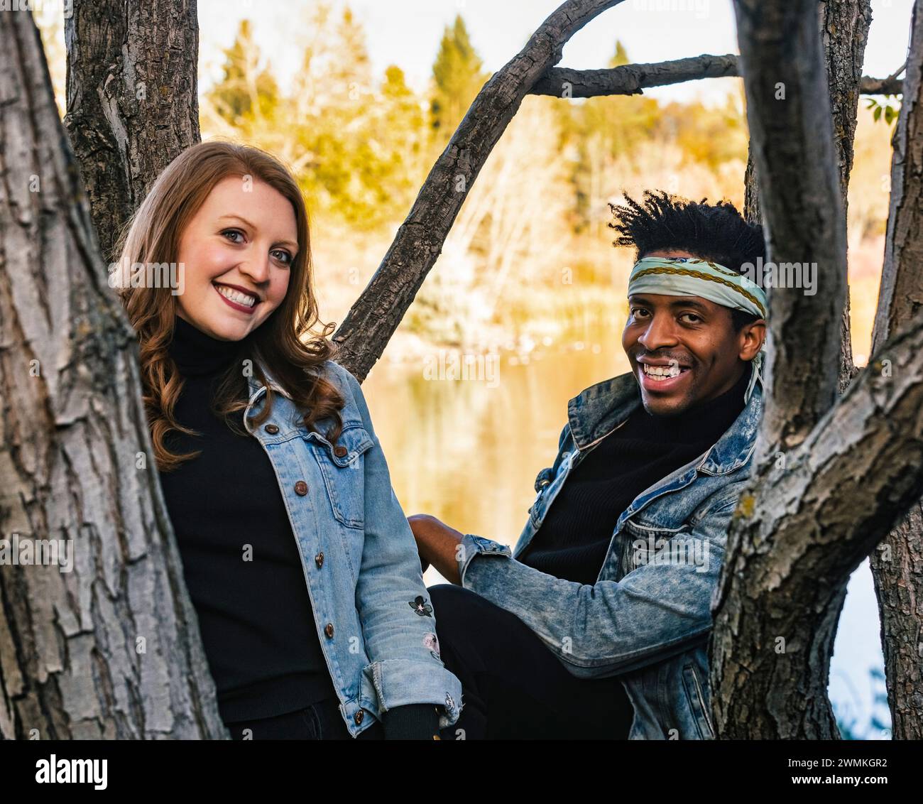 Ritratto ravvicinato di una coppia mista che sorride e guarda la telecamera tra i rami degli alberi, trascorrendo del tempo insieme durante un fami autunnale... Foto Stock