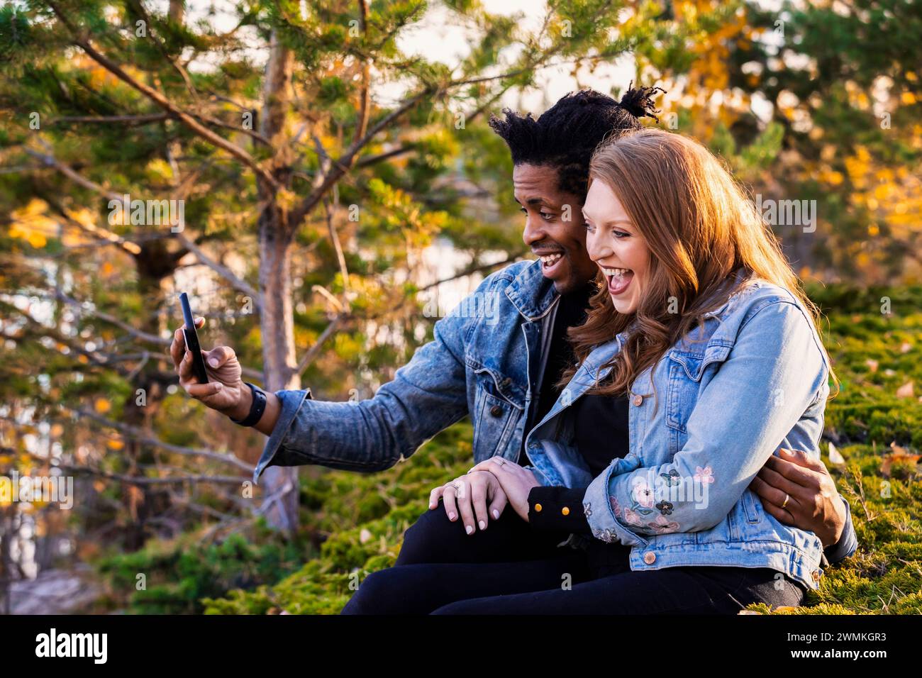 Primo piano di una coppia di sposi che sorride e si fa un selfie insieme, seduto sull'erba al crepuscolo, trascorrendo del tempo insieme... Foto Stock