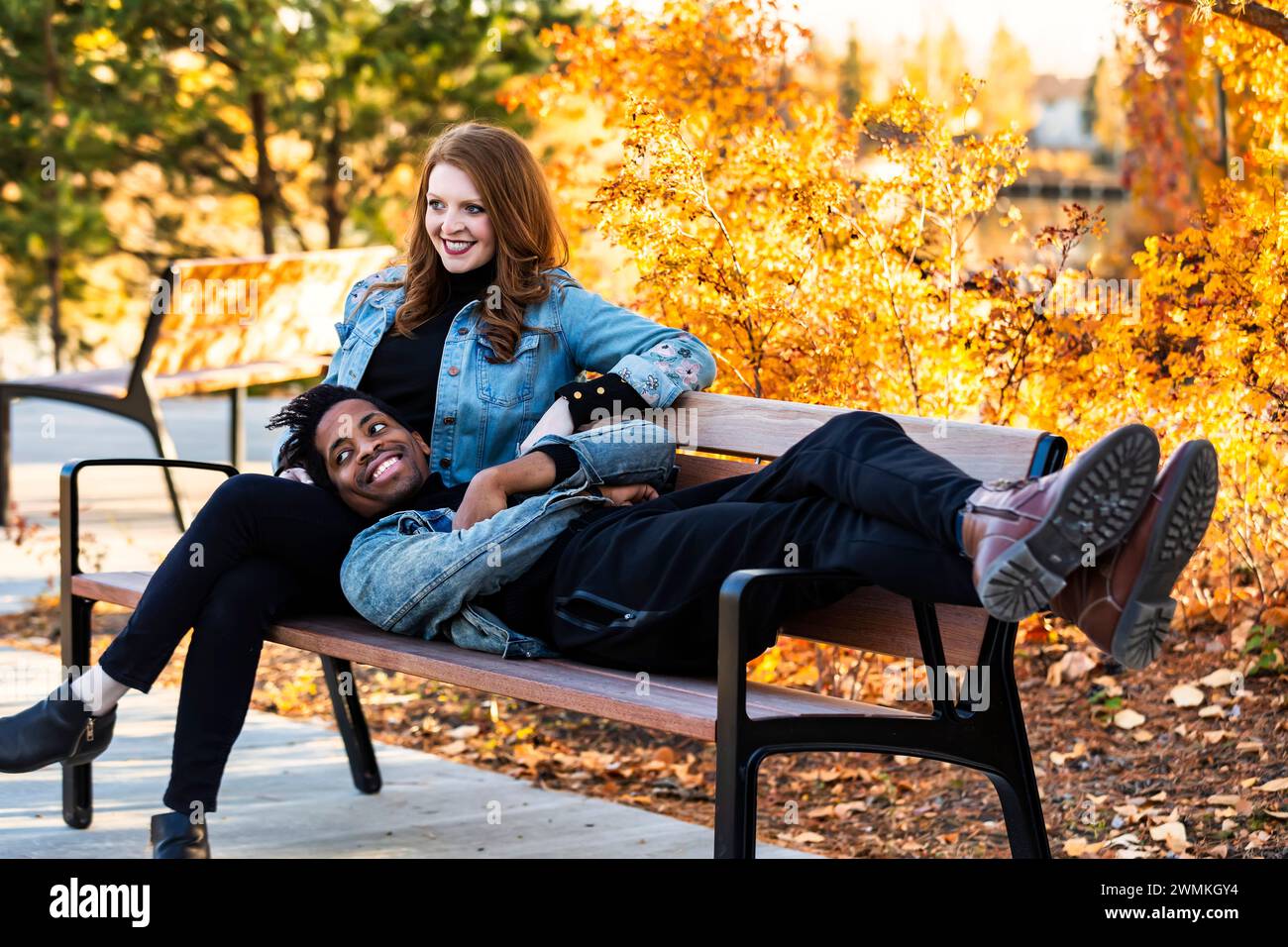 Una coppia di corse miste sorridente e rilassante su una panchina del parco mentre trascorri del tempo insieme durante una gita in famiglia autunnale in un parco cittadino Foto Stock