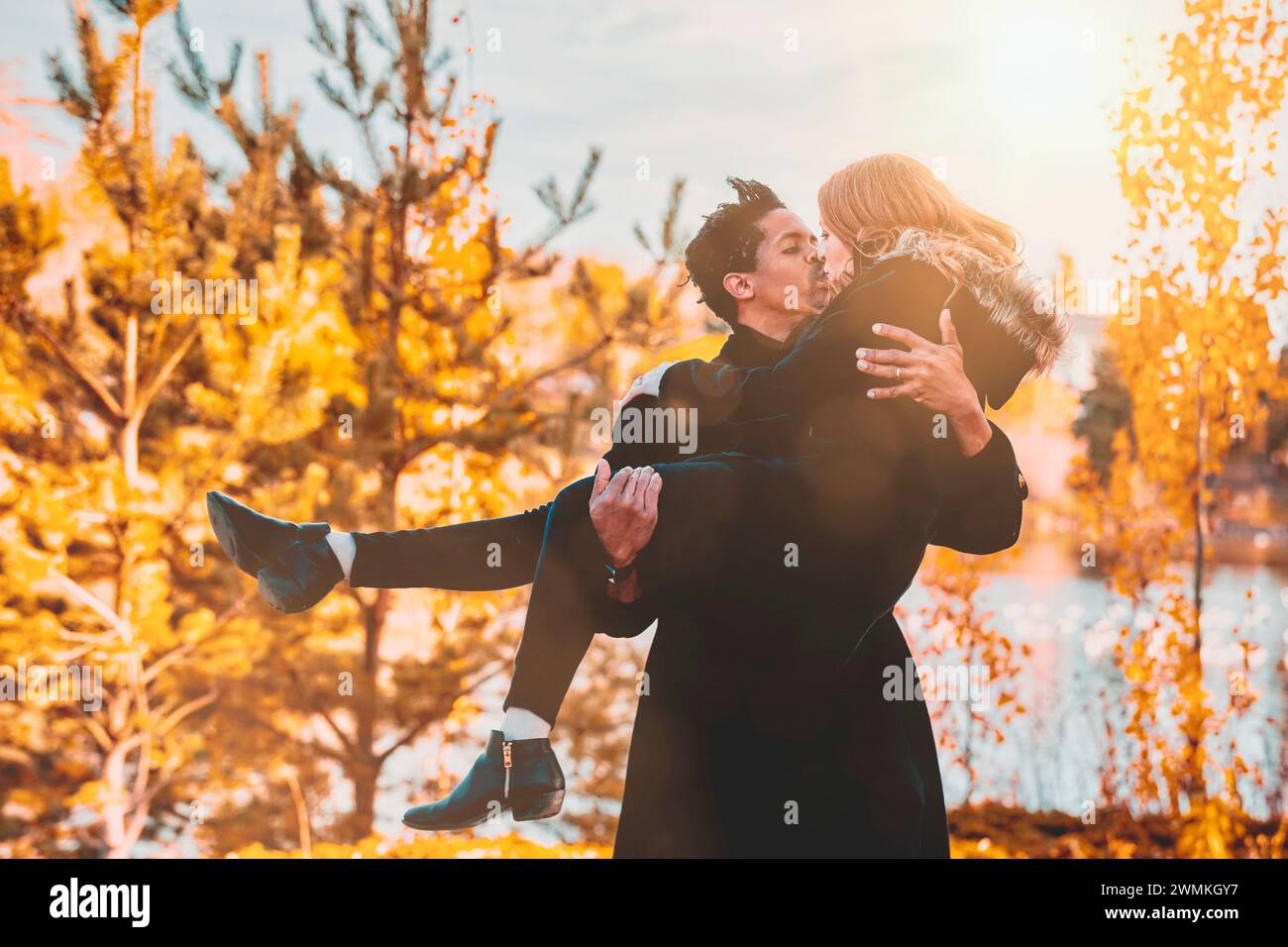 Una coppia mista, marito che porta e tiene in braccio sua moglie mentre condividono un bacio e trascorrono del tempo insieme durante una famiglia autunnale... Foto Stock