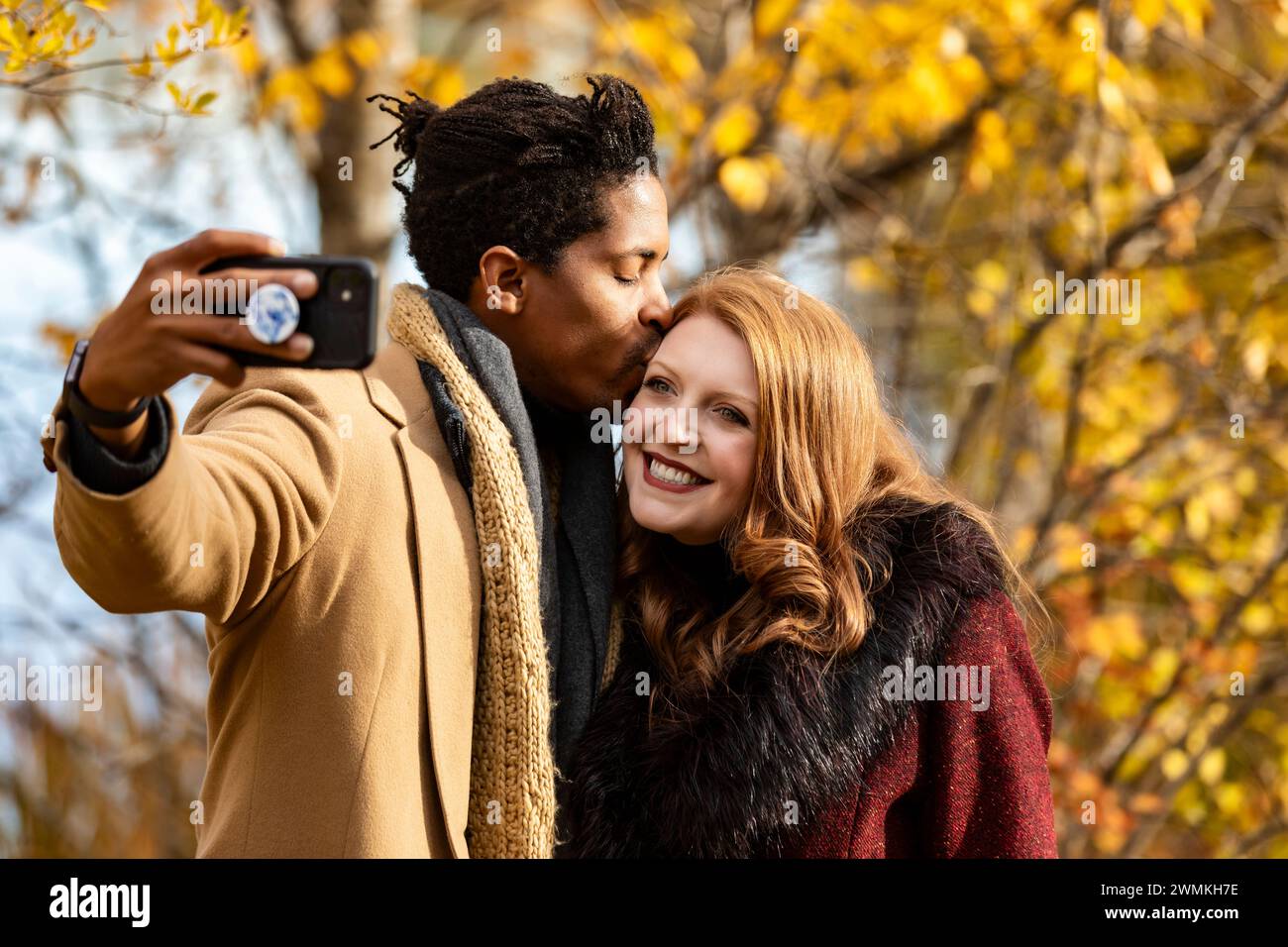 Primo piano di una coppia mista che fa un selfie insieme, marito che bacia la moglie sulla fronte mentre sorride alla telecamera del cellulare, mentre spende q.. Foto Stock