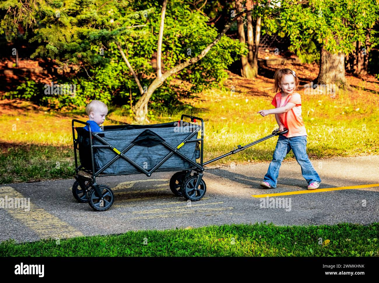 Ragazza in età prescolare che tira suo fratello minore che ha la sindrome di Down in un carro su un sentiero in un parco cittadino durante una calda serata autunnale Foto Stock
