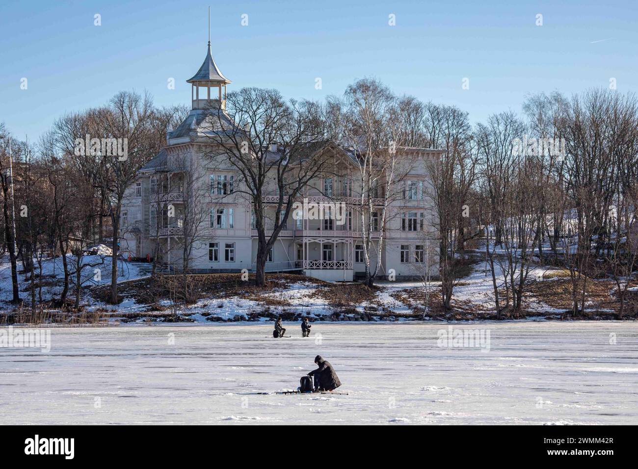 Pesca ghiacciata nella baia di Töölönlahti ghiacciata con una delle ville di Linnunlaulu sullo sfondo a Helsinki, Finlandia Foto Stock