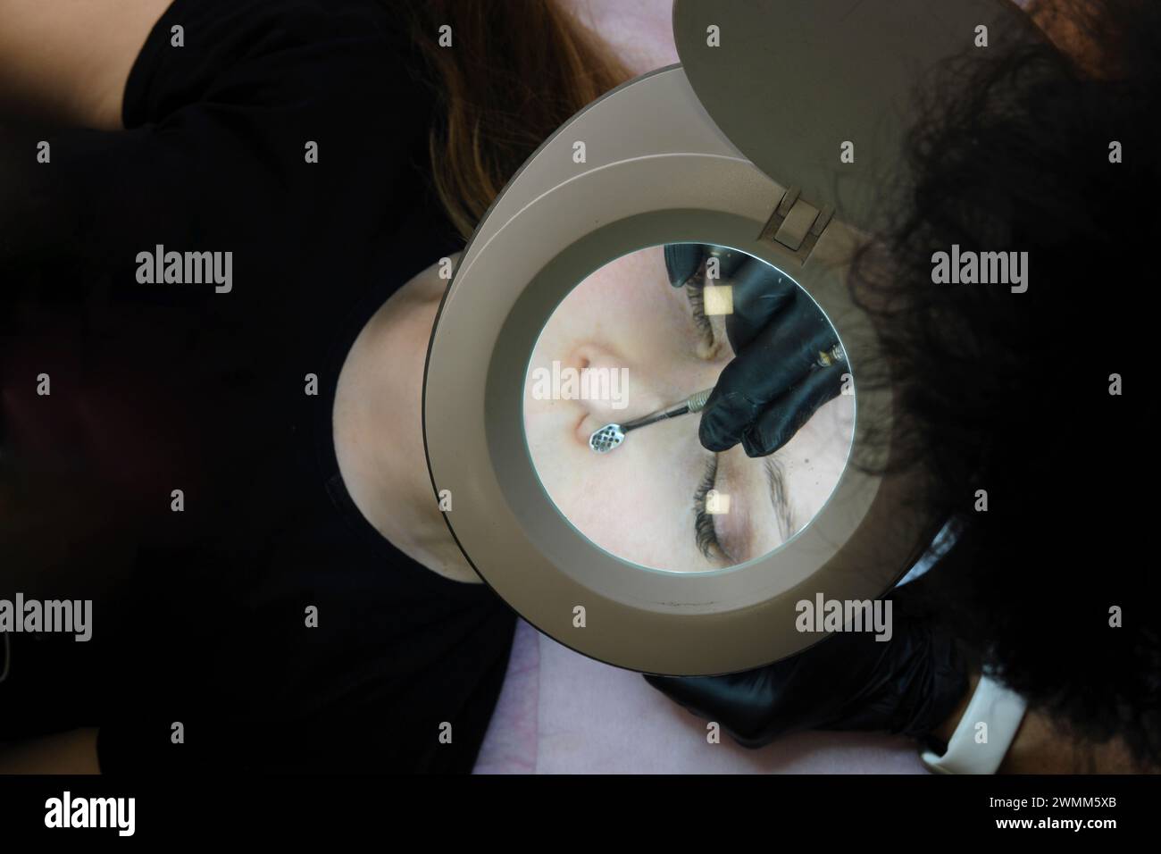 Un cosmetologo durante una sessione di pulizia del viso utilizzando attrezzature speciali per trattare la pelle del cliente, osserva attraverso una lampada con lente d'ingrandimento. Foto orizzontale Foto Stock