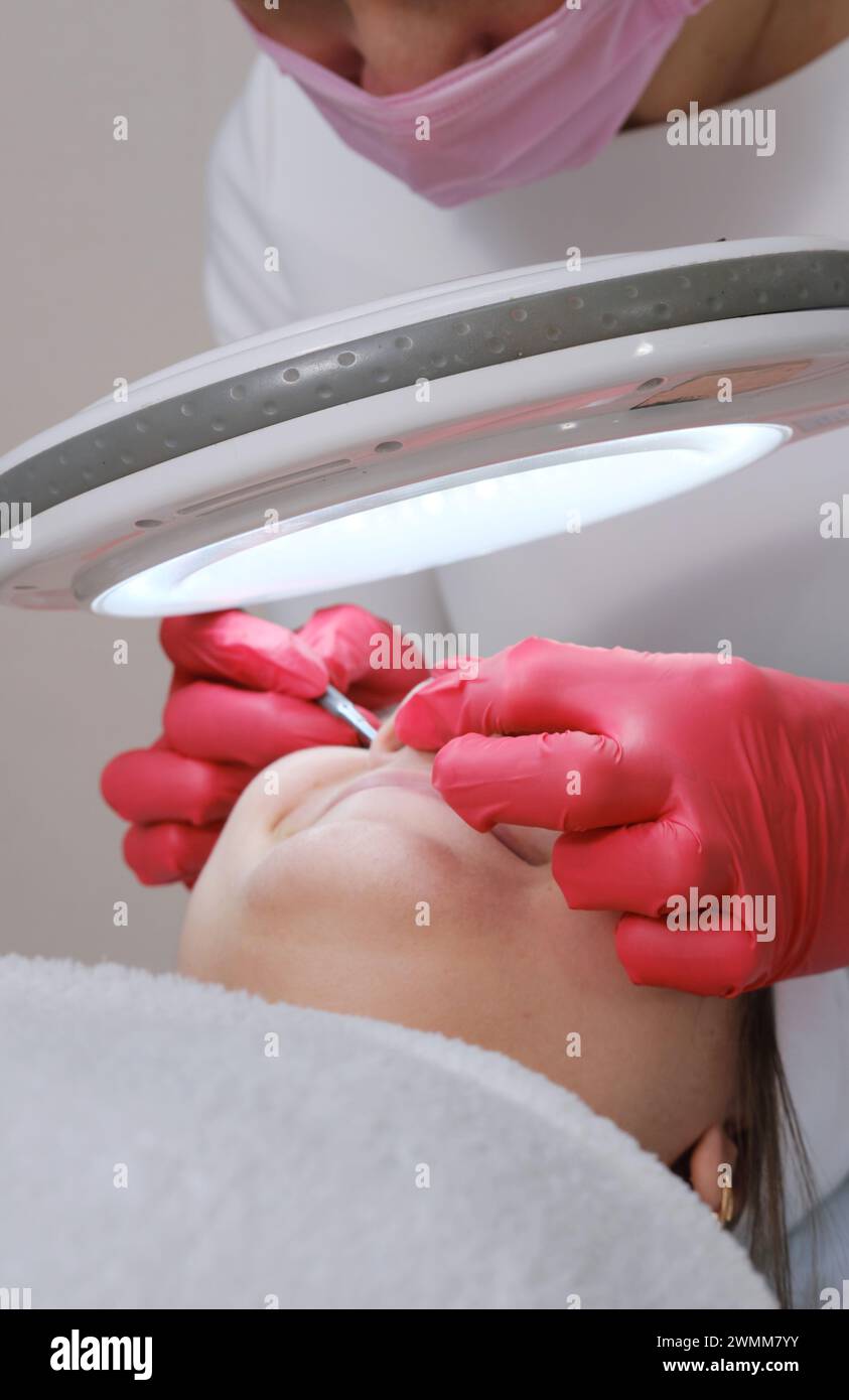 Un cosmetologo con maschera rosa esegue una pulizia del viso su un cliente utilizzando uno strumento speciale sotto la luce brillante di una lampada con lente d'ingrandimento Foto Stock