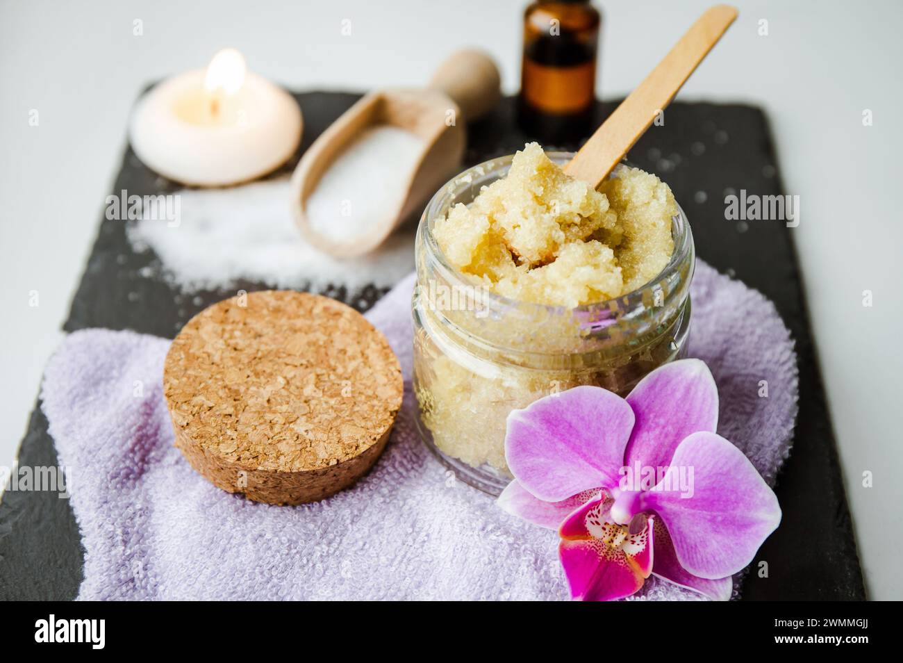 Scrub corpo di zucchero fatto a mano nel vaso di vetro. Candele accese e fiori di orchidea rosa per decorazioni su sfondo bianco in pietra nera. Foto Stock