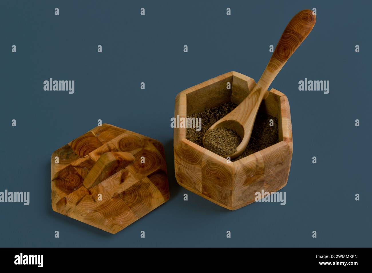 Immagine di natura morta minimalista di una scatola di legno a sei lati contenente caffè macinato con un cucchiaio di legno e un coperchio di legno su un semplice sfondo grigio Foto Stock