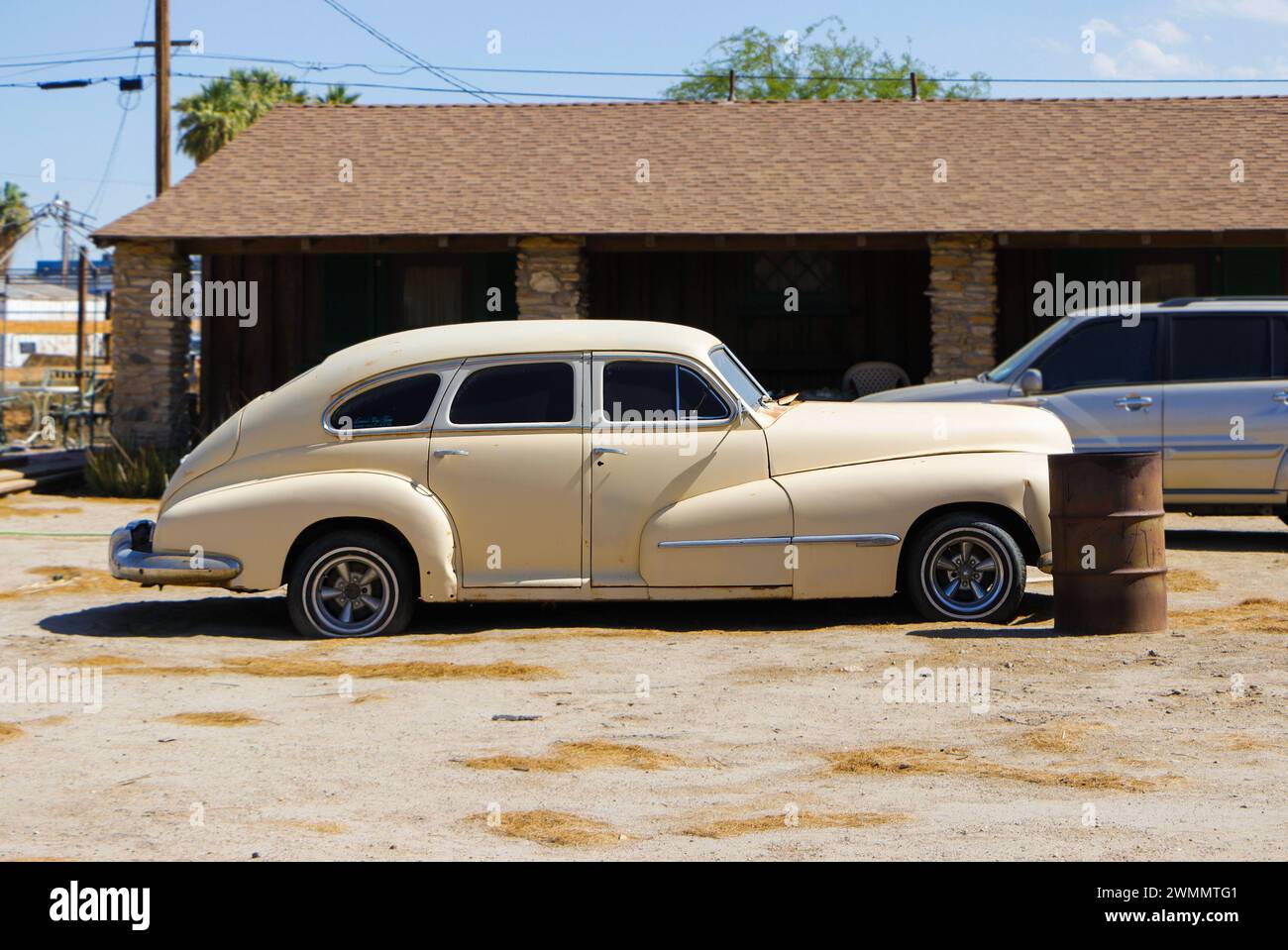 Un'auto beige d'epoca parcheggiata fuori in un'area desertica accanto a un vecchio edificio e a un tamburo di olio. Foto Stock