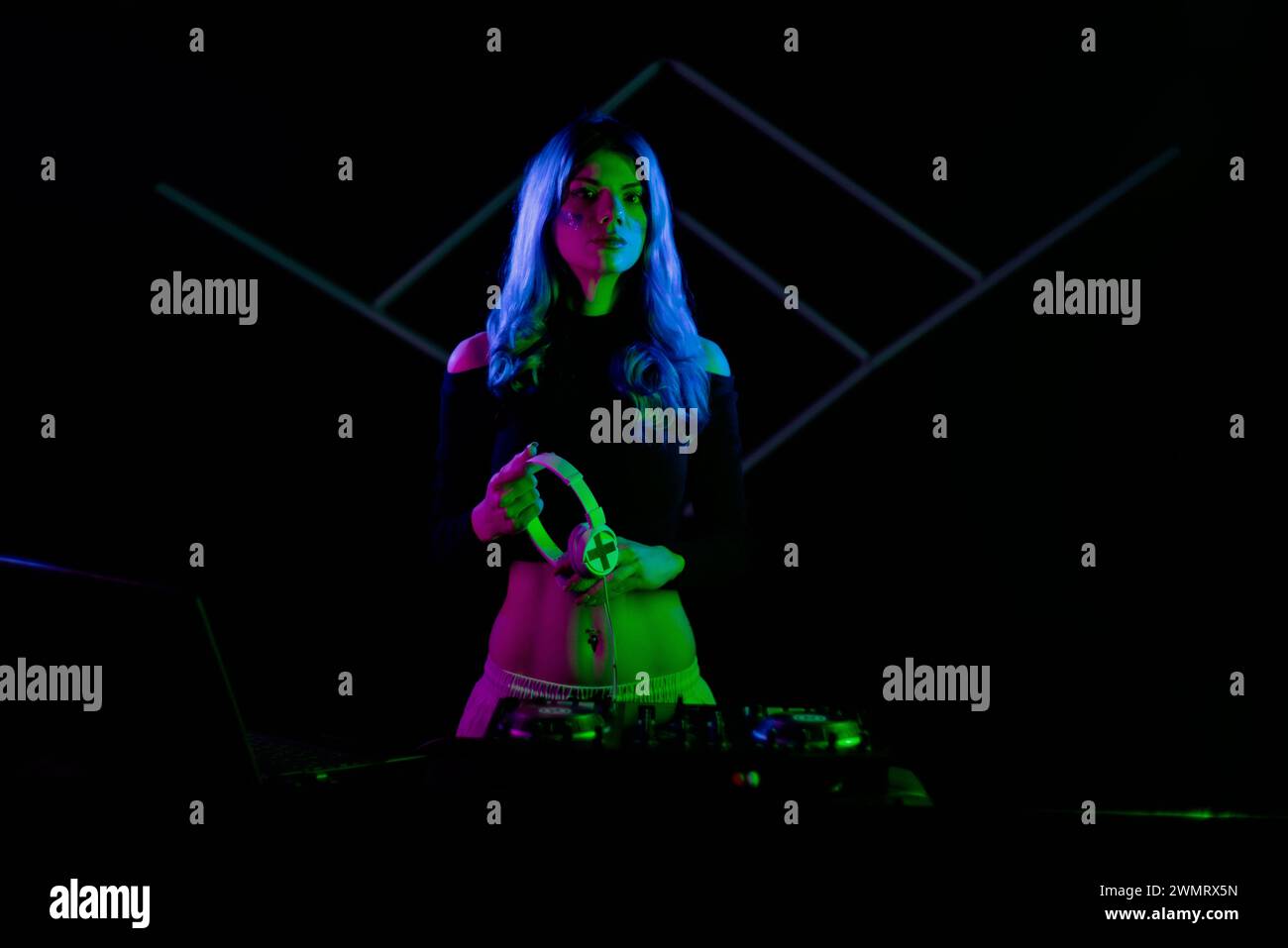 Una donna con i capelli blu vibranti sta facendo il DJ su un giradischi. Tenere le cuffie e guardare la fotocamera. Illuminata lateralmente con luci al neon colorate Foto Stock