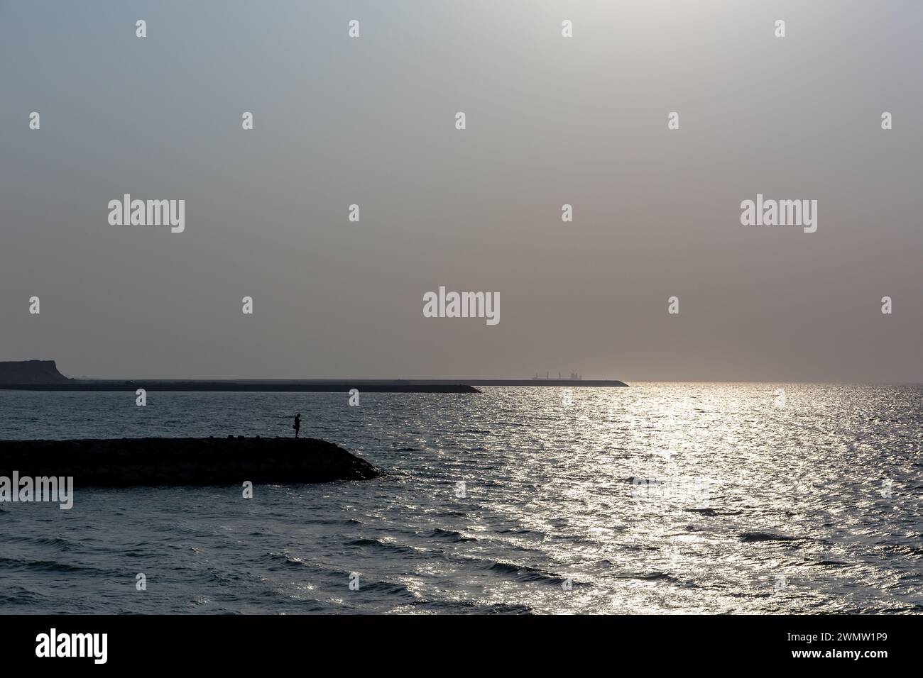 Sagome di frangiflutti di pietra con una persona in piedi alla fine con una dura luce solare che si riflette nelle onde del Golfo, la spiaggia di al Hamriyah negli Emirati Arabi Uniti. Foto Stock