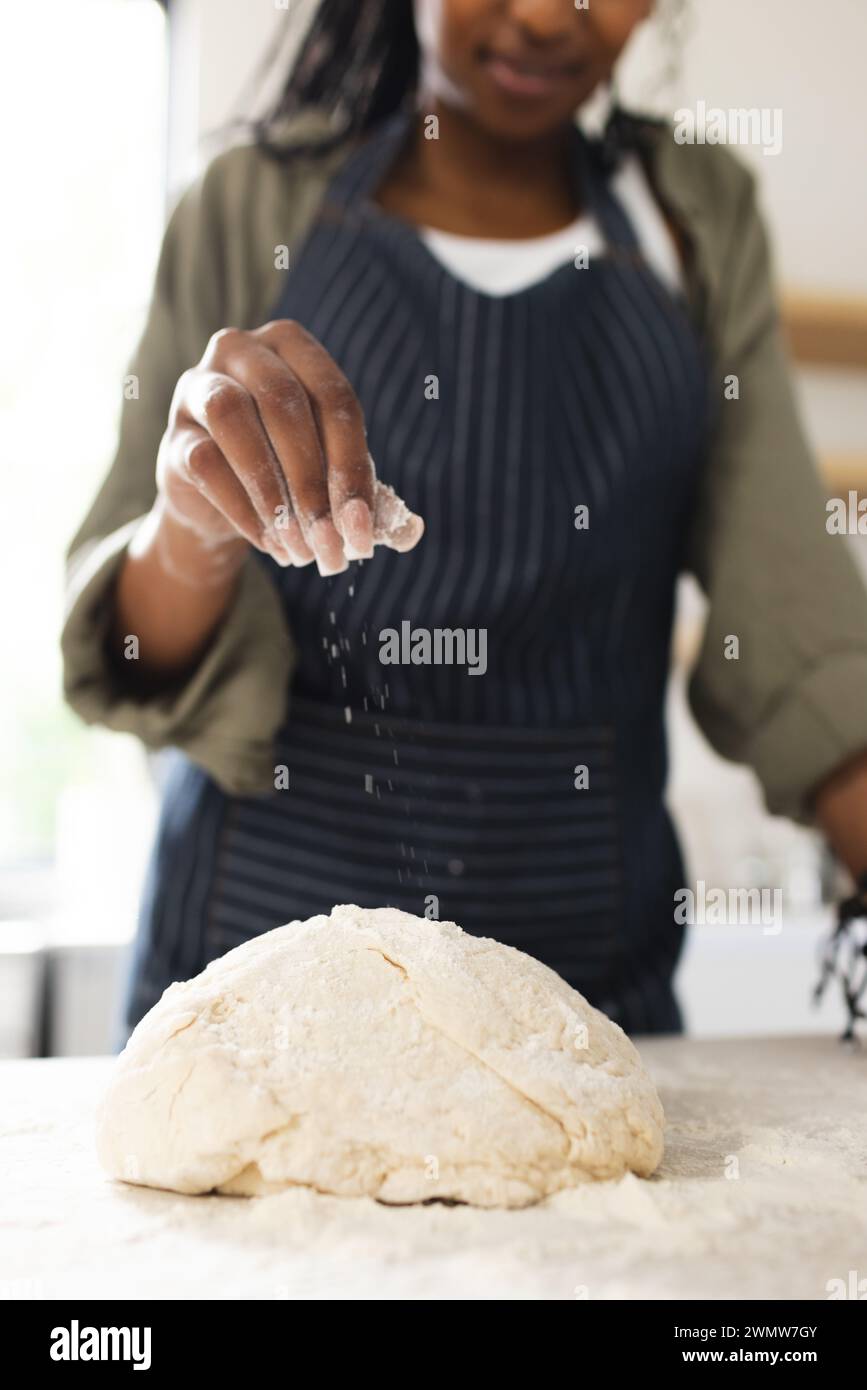 Una giovane donna afroamericana spruzza la farina sull'impasto su un bancone della cucina con spazio per le copie Foto Stock