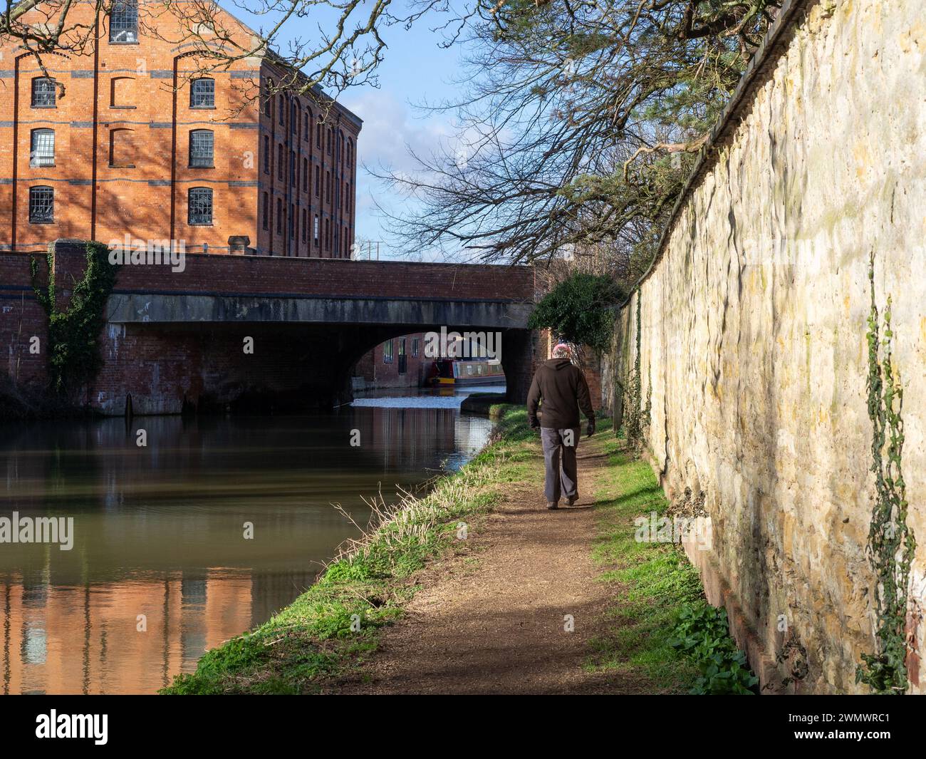 Vista posteriore di un uomo che cammina lungo un sentiero lungo il canale Grand Union durante il sole invernale, Blisworth, Northamptonshire, Regno Unito; Blisworth Mill sul retro Foto Stock