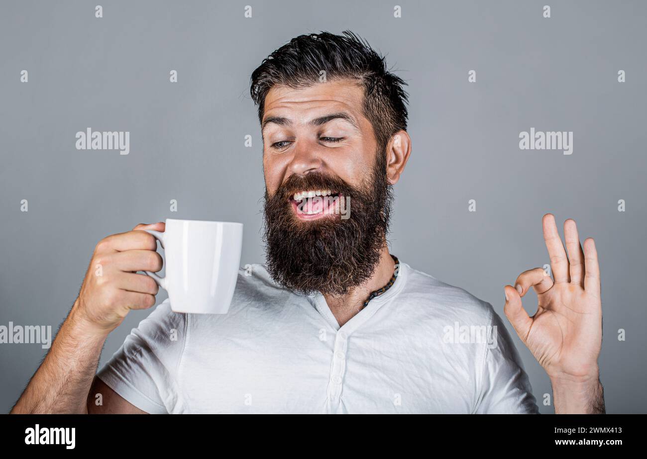 Uomo sorridente felice che tiene in mano una tazza di gustoso caffè fresco e mostra il pollice in su. L'uomo regge una tazza di caffè, tè. Uomo barbuto sorridente mostrando un segno ok. Bene Foto Stock