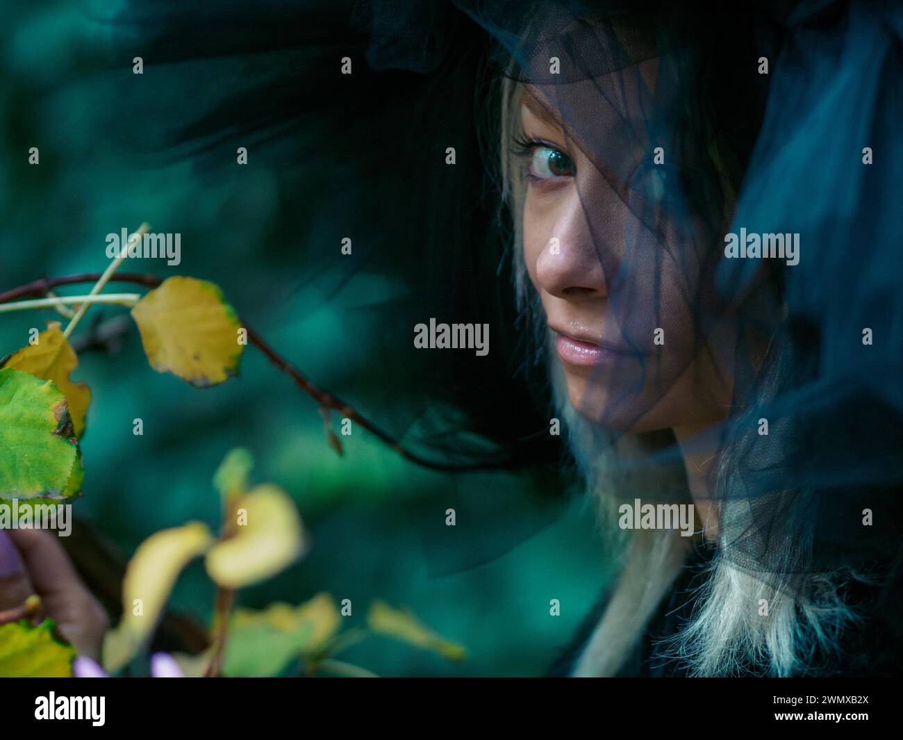 Lo sguardo intenso di una giovane donna bionda caucasica sottile e pallida attraverso le foglie, che indossa un sostanzioso cappello nero in tulle, suggerendo un pensiero profondo Foto Stock