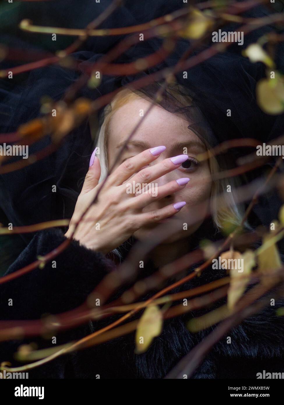 Immagine vivida di una giovane donna bianca pallida che sbircia attraverso le foglie d'autunno con la mano che copre parte del suo viso indossa un velo di pizzo Foto Stock