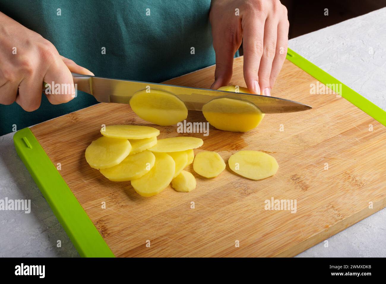 Autentiche mani femminili che tagliano patate su un tagliere di legno sul tavolo della cucina. Donna in grembiule ha tagliato patate, preparando cibo, interni domestici, l Foto Stock