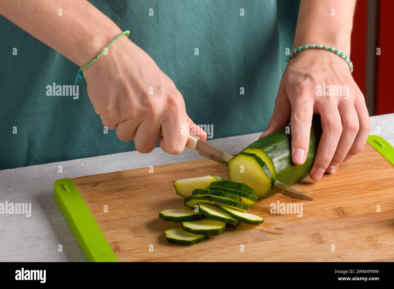 Autentiche mani femminili che tagliano zucchine su un tagliere in legno sul tavolo della cucina. La donna in grembiule tagliò le zucchine per preparare il cibo. Vegetariano e vegetariano Foto Stock